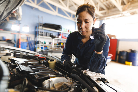 Mixed race female car mechanic wearing overalls, inspecting car and using torch
