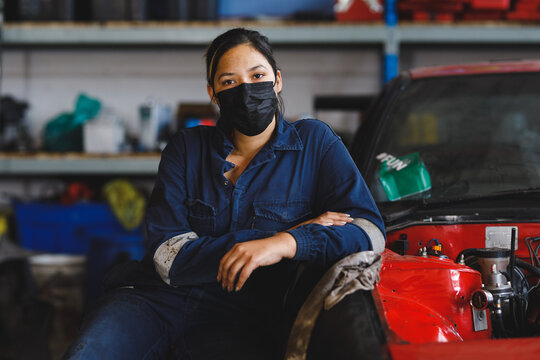Mixed race female car mechanic wearing face mask and overalls, looking at camera
