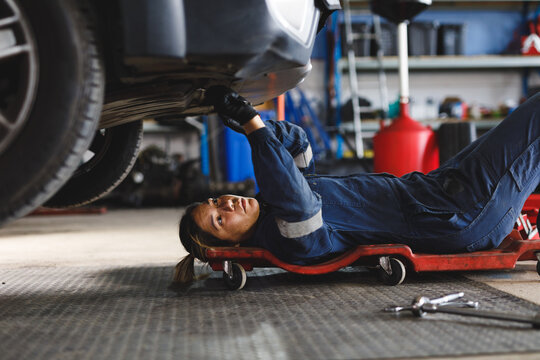 Mixed race female car mechanic wearing overalls, lying on a board, checking car