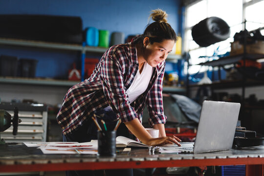 Mixed race female car mechanic standing at desk, using laptop in workshop
