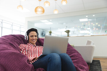 Smiling caucasian female business creative wearing headphones, lying on sofa and using laptop