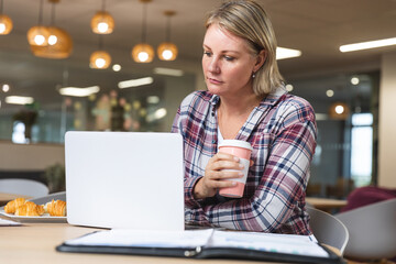 Thoughtful caucasian female business creative holding coffee and using laptop at workplace cafeteria