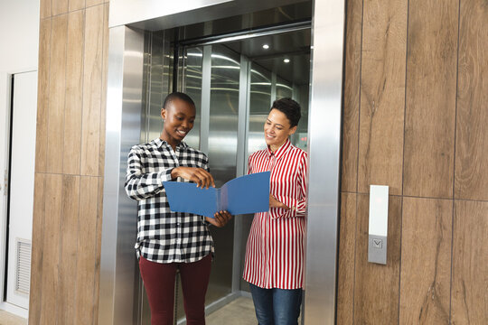 Happy diverse female creative colleagues holding notes and talking in workplace lounge area