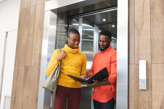 Smiling african american male and female creative colleagues holding notes, talking at elevator