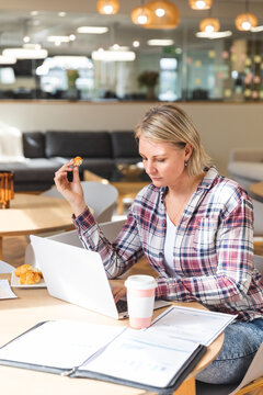 Thoughtful caucasian female business creative holding cookie and using laptop at workplace cafeteria