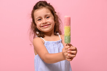 Funny cheerful little girl smiles showing to the camera a delicious sweet frozen juice, popsicle, ice cream on stick in her hands. Summer concept, pink background, copy space
