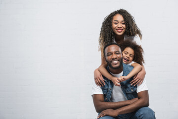 cheerful african american woman with daughter embracing happy man on grey