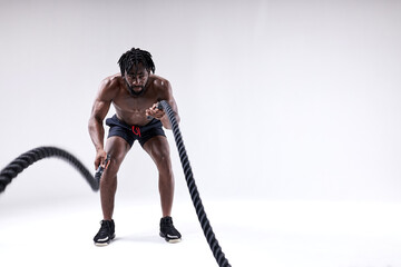 Sporty young black man doing cross fit exercises with battle ropes over isolated white background. hard workout of african shirtless guy. full-length portrait. sport and motivation concept