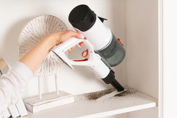 Woman is cleaning shelf in closet with brush of cordless handheld vacuum cleaner. House cleaning, close-up of female hands with vacuum cleaner.