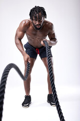 Fototapeta na wymiar Full-length Portrait Of Young African American Guy With Naked Torso Exercising With Battle Ropes, Cross Fit Training Isolated On White Background. Confident Concentrated Man Is Keeping Fit. Sport