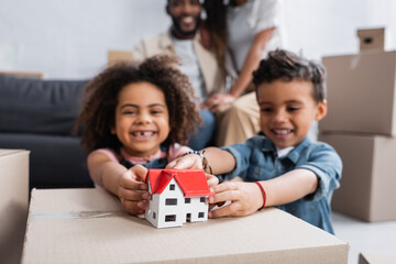 cheerful african american kids near house model and parents on blurred background