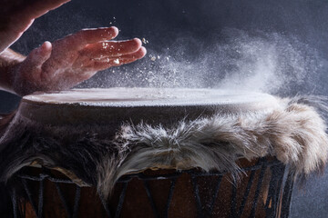 Man is playing on djembe drum, covered with talcum powder. Flour splashes on dark background....