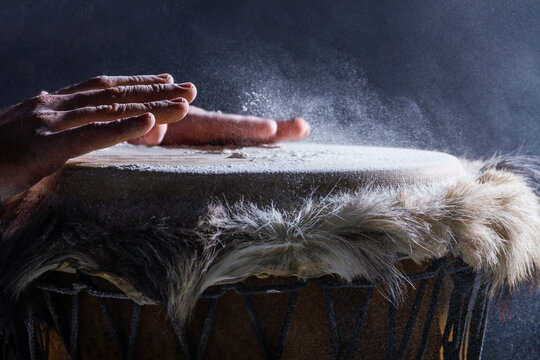 Man is playing on djembe drum, covered with talcum powder. Flour splashes on dark background. Summer festival concert performance. Ethnic rhythm. Percussion musical instruments and culture concept.