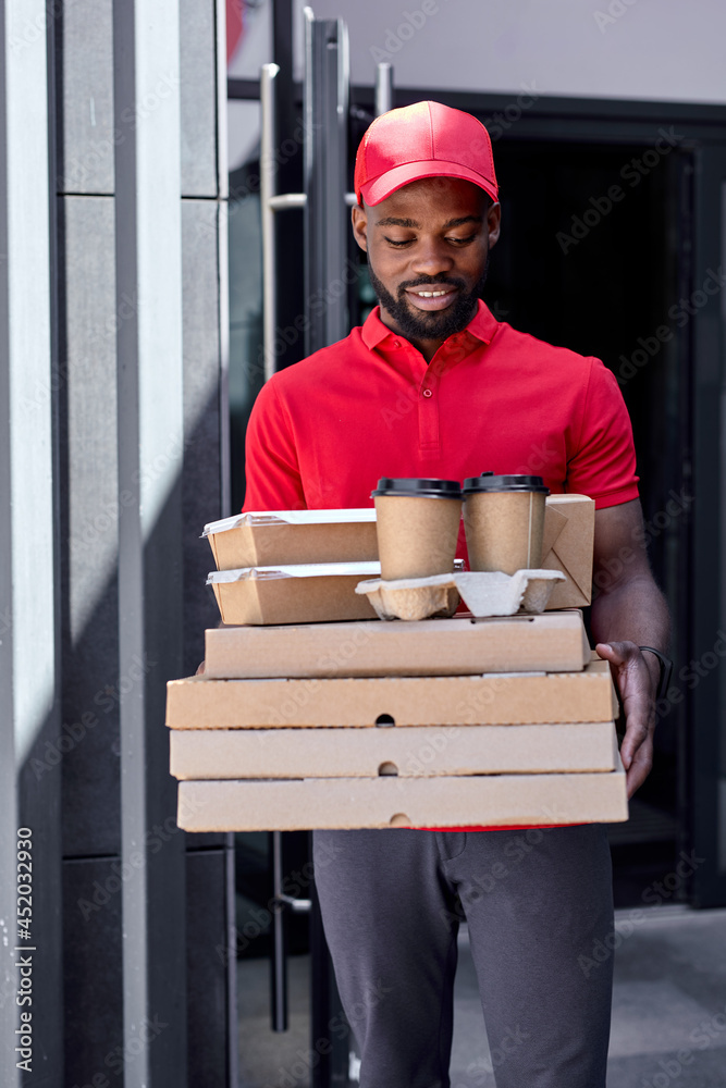 Wall mural Handsome black afro courier dilivering food, drinks to clients customers. Young man in red uniform is carrying order. Food delivery in quarantine. Courier bring Pizza and coffee for customer