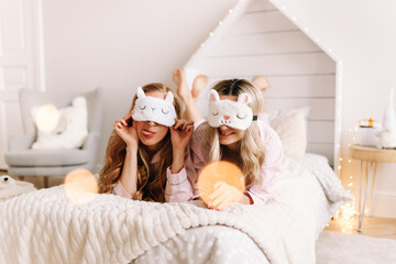 Obraz na płótnie Canvas Pretty funny two young women in pajamas and sleep masks having fun laughing relaxing lying on the bed in a cozy decorated bedroom at home during the Christmas holidays, selective focus