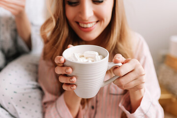 Hot winter drinks. Happy women in pajamas hold mugs of coffee with marshmallows in a cozy bedroom at home in winter, a selective focus