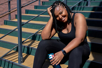 Obraz na płótnie Canvas African Exhausted Female In Black Sportive Clothes Have Rest After Sport, Sitting With Water Bottle, Having Rest, Looking At Camera Smiling, Posing. In The Morning, Weight Loss, Sport, Fitness