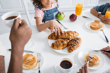 cropped view of african american girl taking croissant during breakfast with blurred family