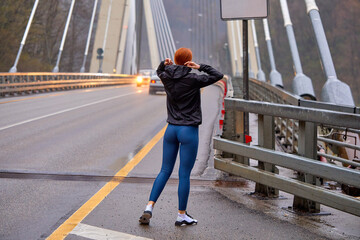 Rear view on young female in sportswear walking on bridge having rest after running workout, at cold spring day, redhead slim female outdoors, healthy active lifestyle, sport and fitness concept