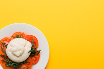Delicious fresh cheese made from cream and milk - burrata. Italian tender cheese with tomato and greens salad on a bright yellow background