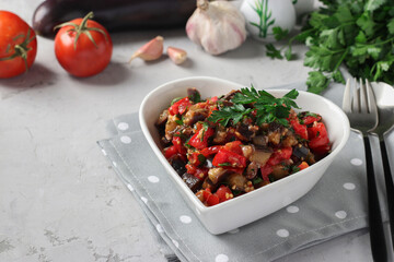 Salad with eggplants, tomatoes, garlic and parsley in a plate in the shape of a heart on a light gray background.