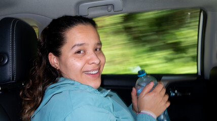 joven mujer a bordo de auto con botella de agua en la mano sonrie a la cámara con fondo barrido detrás de ella 
