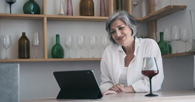 Mature Elderly 60 Year Old Woman Standing With A Tablet, Drinking Wine And Enjoying A Remote Conference With Friends, Enjoying A Family Distant Video Chat Celebration, Active Leisure Feeling Alone