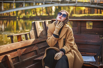 Elegant young lady in long coat with magazine on wooden boat on the river in autumn park