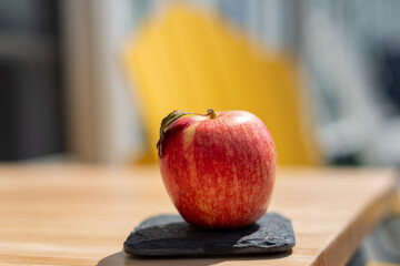 A red, gala, apple sits on a small square grey slate on a wooden table. The sweet crisp fruit has a...