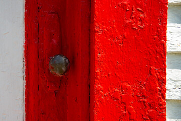 A closeup of an antique rusty iron decorative metal door handle on a brightly painted vibrant red wooden door.  The vintage door is next to a white wall and glass window that's paint is peeling. 