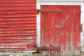 Obraz na płótnie Canvas A vibrant vintage red wooden barn with a small door, rusty hinges, keyhole, and a latch. There's rot on the wood boards in the bottom of the door. The building has white trim around the door. 