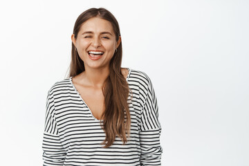 Positive girl winking and showing white healthy smile, sticking tongue playful, standing over white background