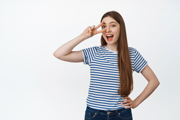 Obraz na płótnie Canvas Cute positive girl show peace, v-sign near eye and laughing, smiling at camera, standing confident against white background