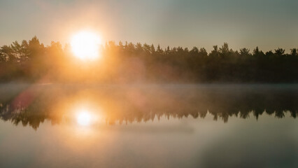 Scenic misty glowing sunrise over lake with large orange Sun, Sun rays in rising from water surface fog coast with trees, mirror reflection in lake surface. Copy space