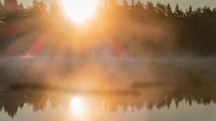 Scenic misty glowing sunrise over lake with large orange Sun, Sun rays in rising from water surface fog coast with trees, mirror reflection in lake surface. Close up photo, copy space