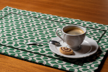 Hot black coffee, served on white crockery, with a small spoon and a two-color biscuit, served over a placemat with a white and green geometric print. With space for text.