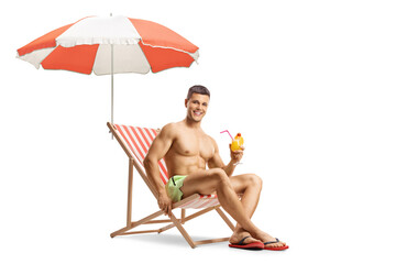 Guy in swimming shorts sitting on a beach chair under umbrella and holding a cocktail