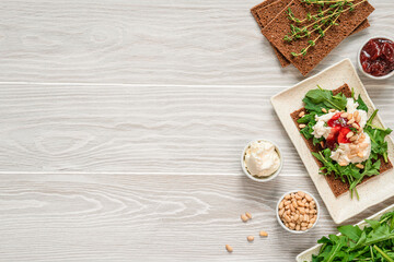 Top view on wooden table with crisp bread and ingredients: cream cheese, jam, arugula and pine nuts. Healthy breakfast concept.