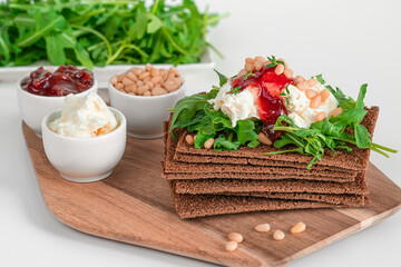 Close-up crisp bread on wooden cutting board and ingredients: cream cheese, jam, arugula and pine nuts. Healthy breakfast concept