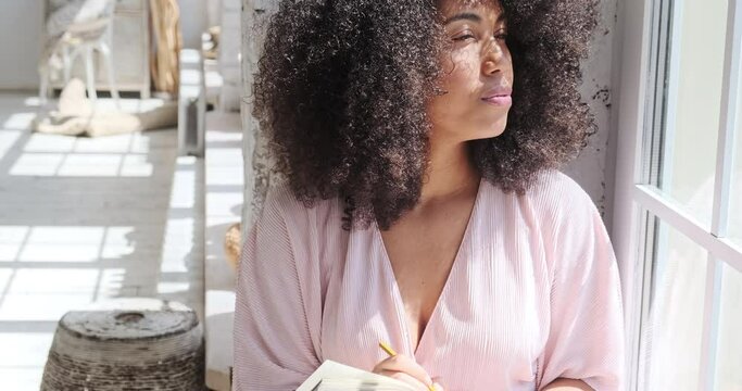 A pensive young black woman with curly hair glances about and writes in a paper notebook near the window. In preparation for exams, the student writes in a notebook and completes assignments.