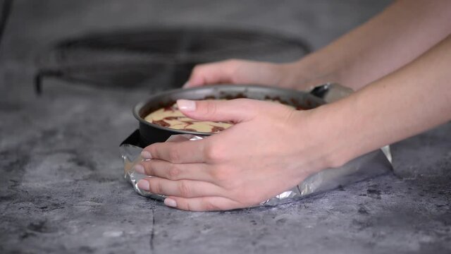 Woman preparing cheesecake with berry sauce in the kitchen. Wrapping the aluminum foil around the springform pan.