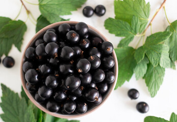 Top view of large, ripe black currant berries in a bowl and leaves on a white background, soft focus.