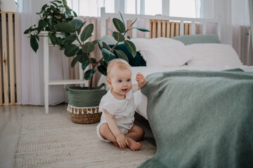 a baby girl in a muslin jumpsuit is sitting on the floor on a rug in the bedroom - obrazy, fototapety, plakaty