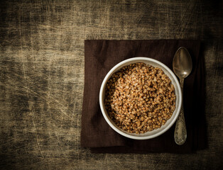 Buckwheat porridge in a white plate and a silver spoon on a dark cloth napkin, placed on a dark wooden background, close up, copy space