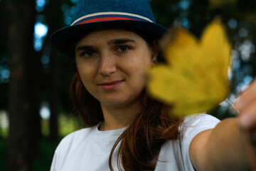 Defocus portrait of a happy young woman with brown hair wearing a hat outdoors. Female hand holding yellow dry leaf. Hello autumn. Women standing at fall park. October mood. Blur. Out of focus