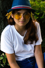 Defocus close-up portrait of a happy dreamy young woman with brown hair wearing a hat with dry yellow leaves and white t-shirt outdoors. Yellow sunglasses. Women seating at trunk tree. Out of focus