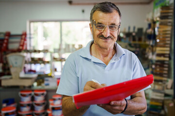 Front view portrait of adult senior caucasian man standing at his store real people small business entrepreneur holding folder with documents checking goods and delivery orders looking to the camera