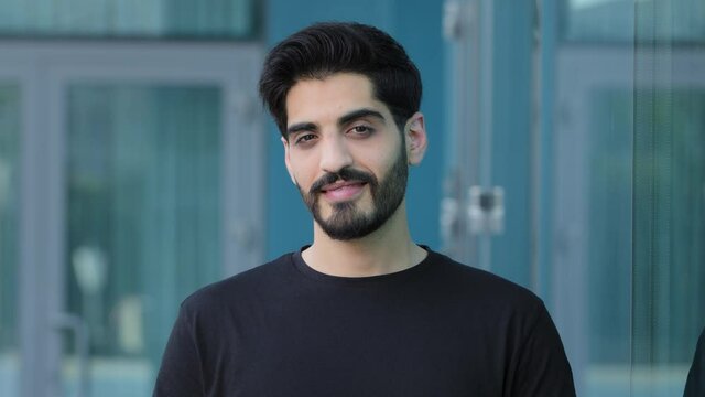 Young Indian Arabic student or businessman with mustache and beard looks affably into camera outdoors. Smiling middle eastern millennial with black hair in summer t-shirt satisfied customer, client