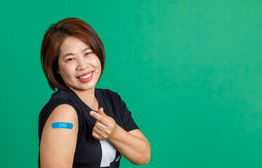 Studio shot of Asian middle aged female patient sit smile look at camera show mini heart hand sign and blue plaster bandage on arm after receive coronavirus covid 19 vaccination on green background