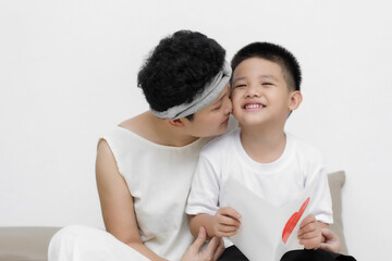 Cute Asian little boy son congratulating his mom happy with Mothers day, giving her handmade greeting postcard with red heart while sitting together on sofa at home. Family holidays concept